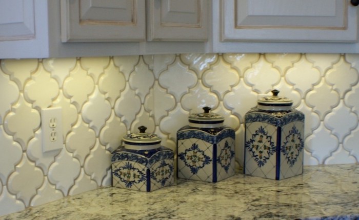 cream-colored arabesque tile backsplash, with a slight 3D effect, near off-white cabinets, and a stone-like, cream and grey counter top, with three porcelain jars