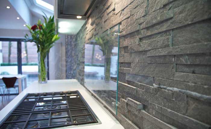 pewter grey stacked stone backsplash, with a clear glass protector, near a white counter top, with a black inbuilt hob