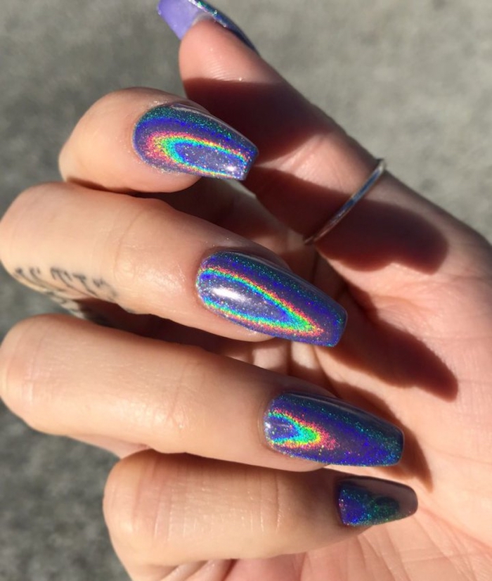 coffin nails covered in iridescent glitter, with hologram effect, and little rainbow reflections, on a hand with a small thumb ring, and a tattoo on the middle finger