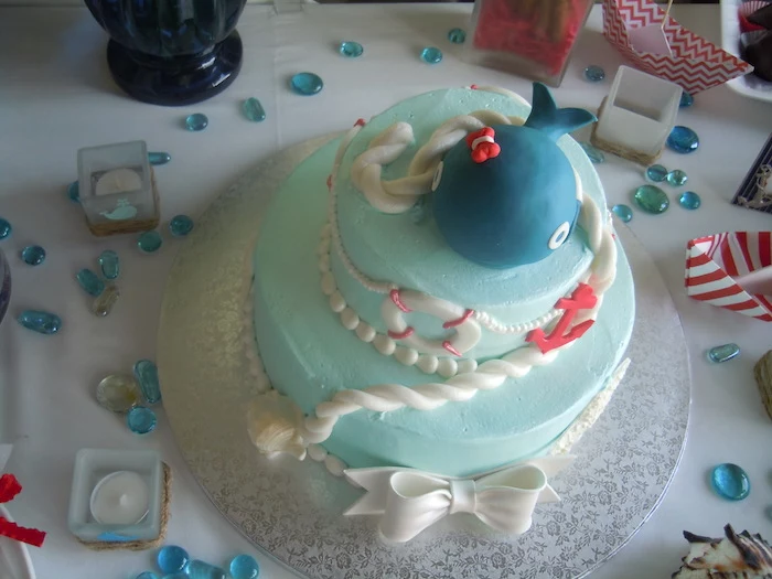 rope and a string of pearls in white, an anchor and a lifebelt, and a dark blue whale figurine, all made from fondant, and topping a creamy pale turquoise cake, nautical baby shower cakes, big white bow