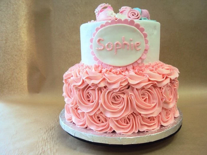 the name sophie written in pink frosting, on a two-layered cake, baby shower cakes for girls, decorated with pink frosted roses, tiny pink fondant shoes and flowers