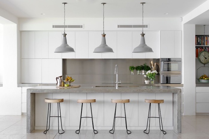 metal back splash, near smooth white cabinets, grey kitchen island, with an inbuilt sink, and four stools, three grey lamps, hanging from the ceiling