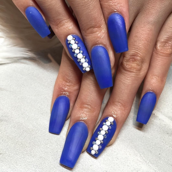 vertical row made from round, white pearl-like nail decal stickers, decorated with smaller iridescent rhinestones, decorating the ring finger nails, of two hands with long acrylic nail shapes, painted in an electric blue color