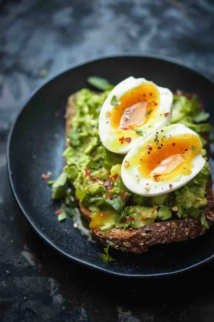 boiled eggs in halves, on top of a piece of bread, with a seasoned guacamole spread, with black pepper an chili flakes, in a black plate