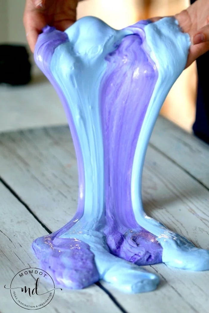 a pair of hands, holding a stretchy piece of slime, in light blue and dark purple, seeping onto a wooden table, in light grey