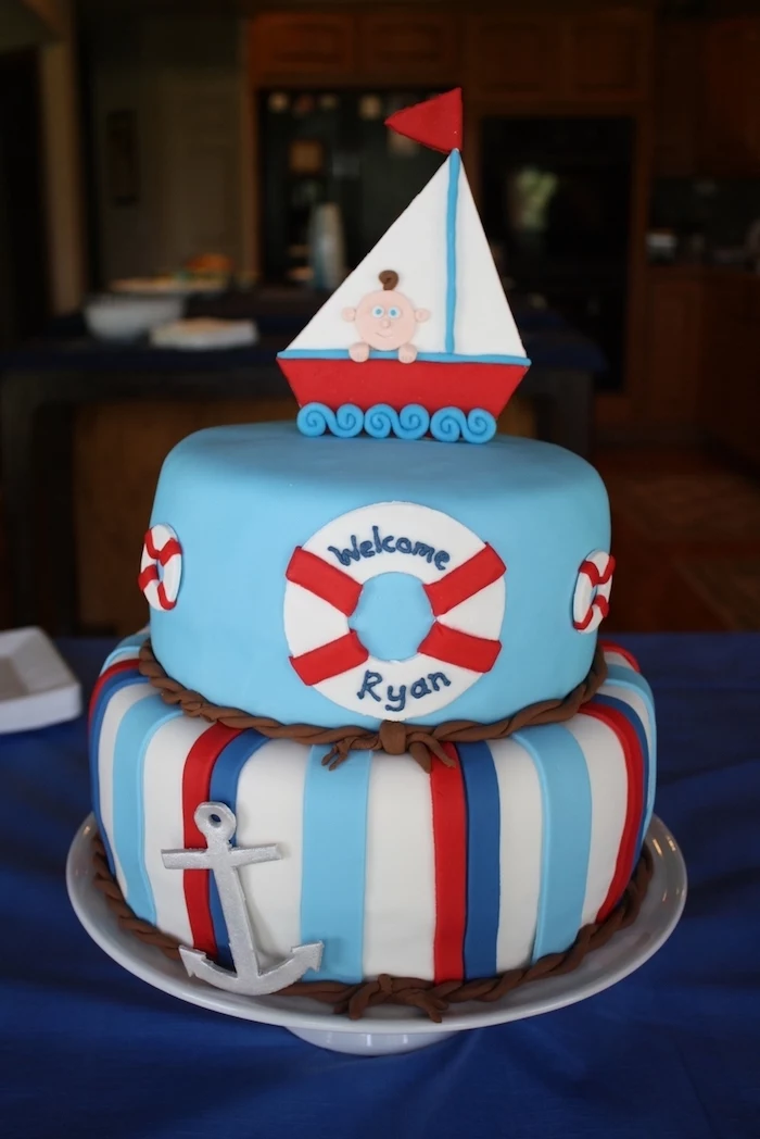 boat made from red, white and blue fondant, and containing a little baby figurine, topping a light blue, two-layered cake, decorated with an anchor and lifebelts