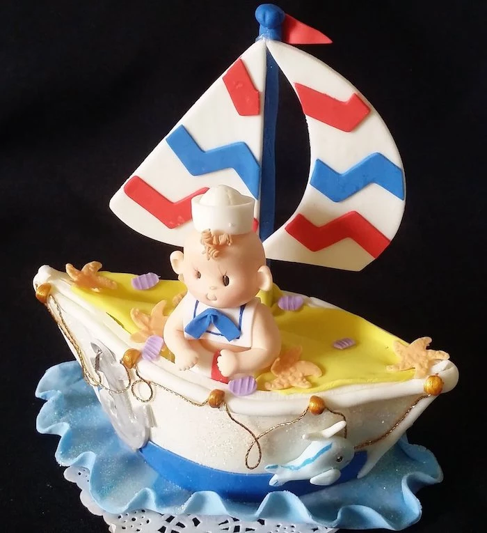 baby shower cake toppers girl or boy, a little fondant sailing boat, in white and red, blue and yellow, containing a smiling baby figurine, in a sailor's outfit