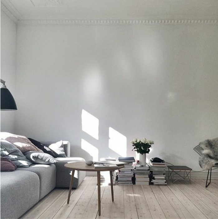books stacked on a light wooden floor, in a minimalistic room, with a pale grey sofa, covered in pastel-colored cushions, three legged coffee table