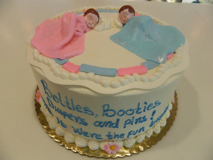 couple of sleeping baby figurines, one covered in a pink blanket, and one in a blue blanket, on top of a white cake, twin baby shower cakes, decorated with a message, done in blue frosting