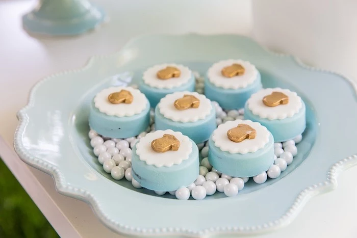 ornate pale blue plate, containing seven pastries, decorated with white, doily-like fondant shapes, and tiny gold-colored baby onesies, onesie cake ideas, white edible pearls 