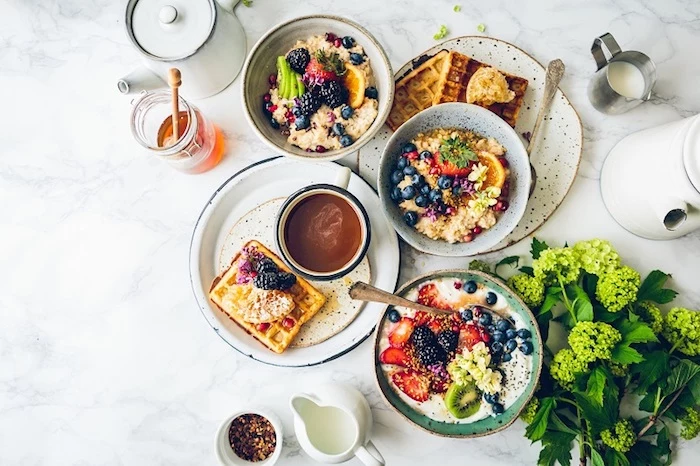 breakfast menu ideas, three bowls of porridge, topped with assorted fruit, placed near or on two plates with waffles, honey and teapots, coffee cup and milk jugs