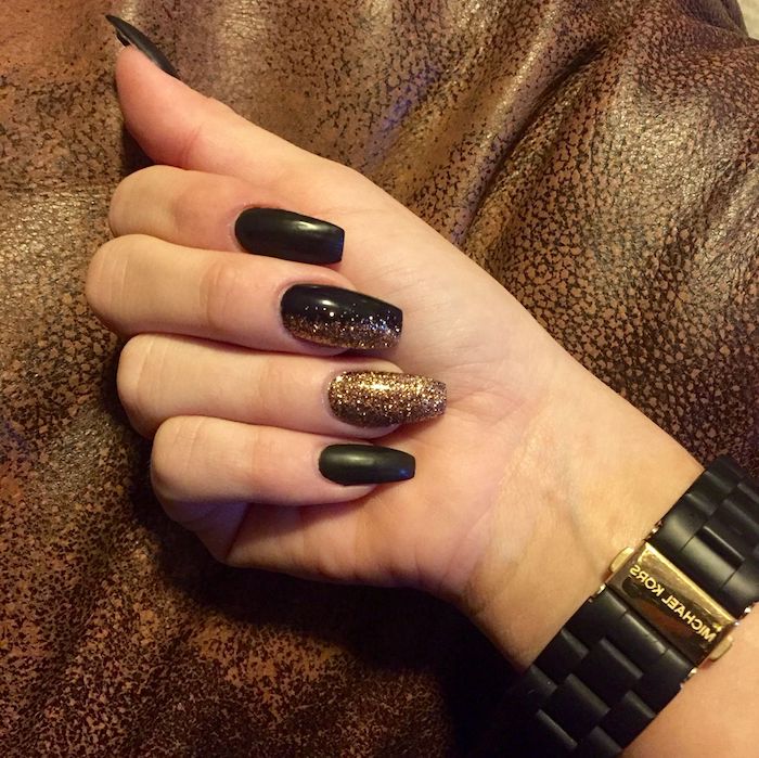 watch in black and gold, on the wrist of a hand with squoval nails, painted in black, and decorated with gold glitter