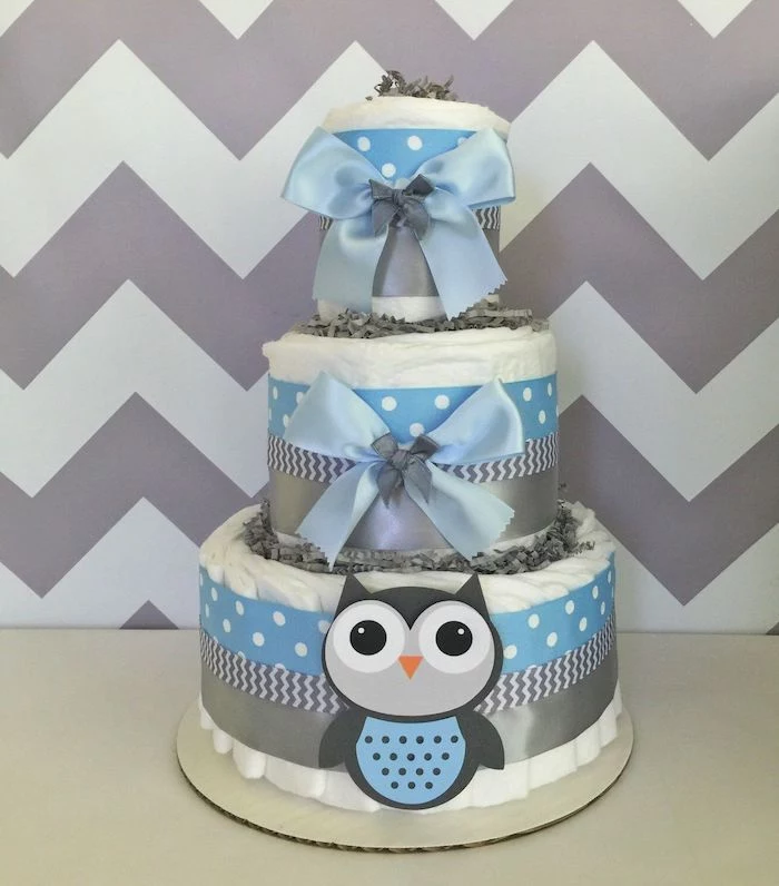 ribbons in pale blue and grey, plain and patterned, on a white diaper cake, with a little owl decoration, owl baby shower cake, grey and white zigzag pattern on the wall behind