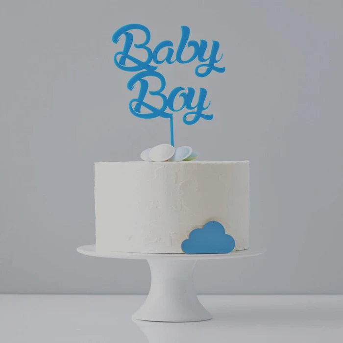 minimalistic cake with white frosting, decorated with several flying saucer candies, and a little blue cloud, on a white cake stand, with a blue topper, reading baby boy