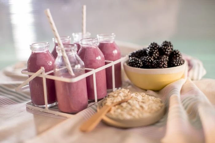 set of six bottles, containing pink smoothies, and two paper straws, on a table near a small bowl of blackberries, and a little plate with rolled oats