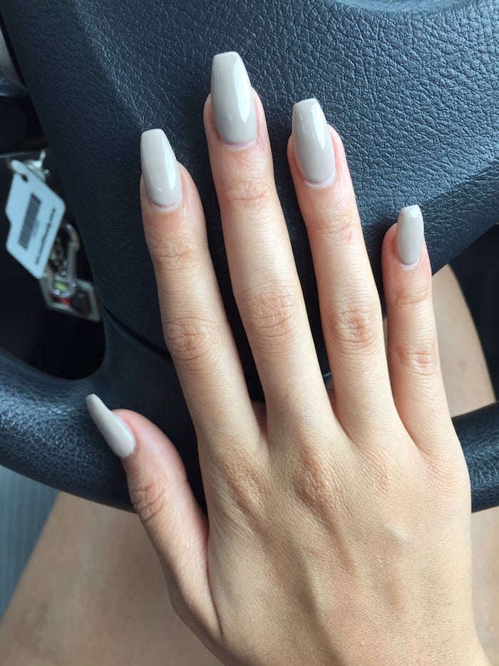 steering wheel in black, under a hand with long slender fingers, and nude coffin nails, in a milky ashen beige hue
