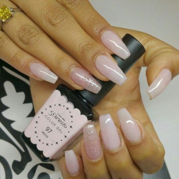 bottle of nail polish, in pale pink and black, held by two hands, with pink nude coffin nails, two of the nails are decorated with pink glitter