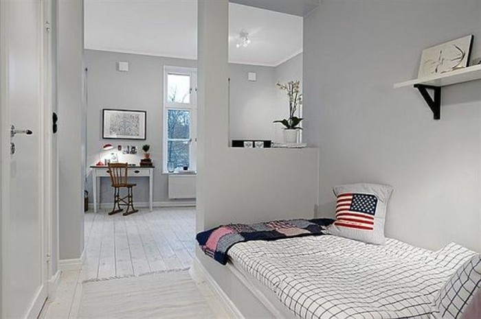 us flag cushion, on a bed with checkered cover, inside a room with pale grey walls, and a single bookshelf, room setup ideas, white desk with a brown chair in the background 