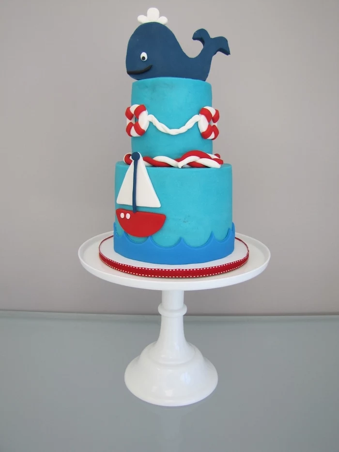 nautical baby shower cakes, smiling whale figurine, made from dark blue fondant, topping a turquoise cake, decorated with red and white details, sailing boat and lifebelts
