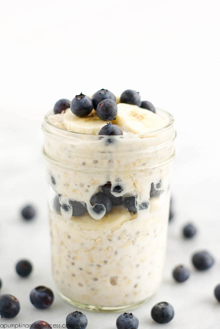 overnight oats served in a glass jar, with blueberries and banana slices, what is a healthy breakfast, on a white surface with other blueberries