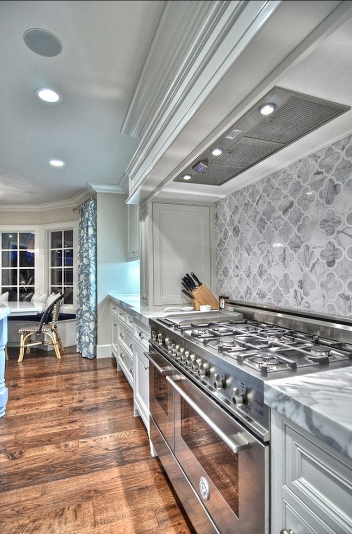 large room with a brown wooden floor, marble counter tops, grey arabesque backsplash, two ovens and off-white kitchen cabinets