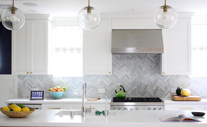 round clear glass lamps, hanging over a white counter top, with an inbuilt sink, in a bright kitchen, with grey herringbone backsplash, and white cabinets