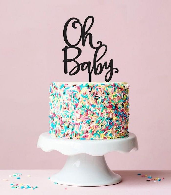multicolored sprinkles covering a tall cake, placed on a white ceramic cake stand, and decorated with a topper, saying oh baby