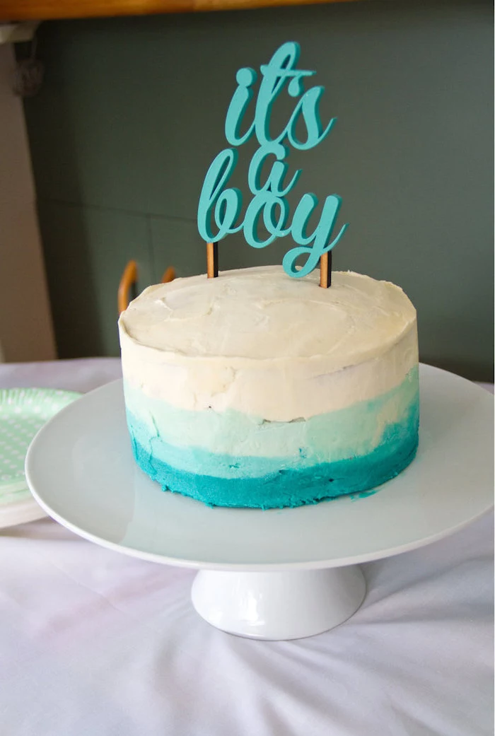 it's a boy written in turquoise, on a topper, decorating a white and turquoise, ombre effect cake, placed on a white ceramic cake stand