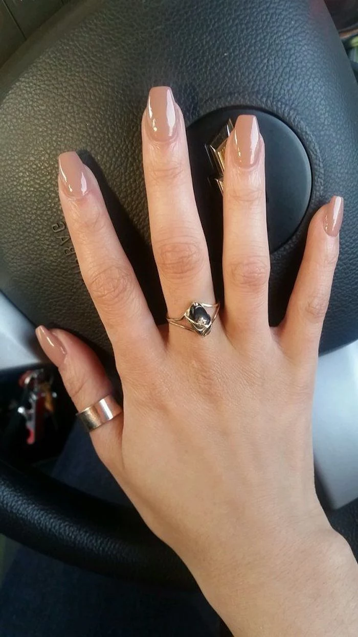 medium length nude coffin nails, on a hand with outstretched fingers, and two golden rings, resting on a black steering wheel of a car
