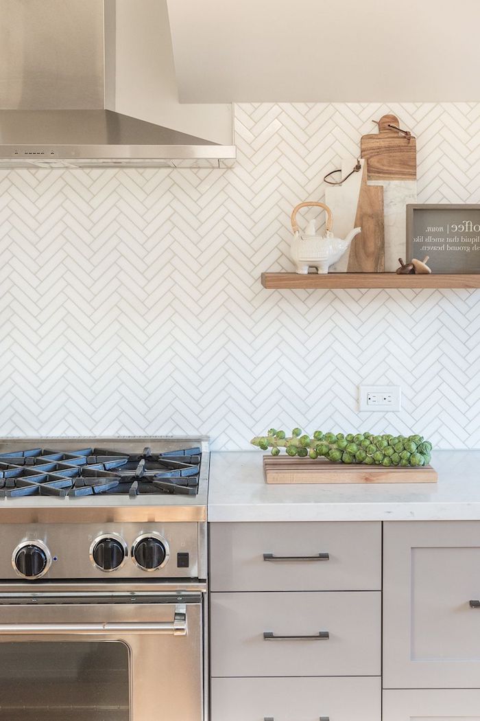 herringbone backsplash in white, with cream-colored joints, near a metal extractor hood, placed over a stove, creamy grey cabinets