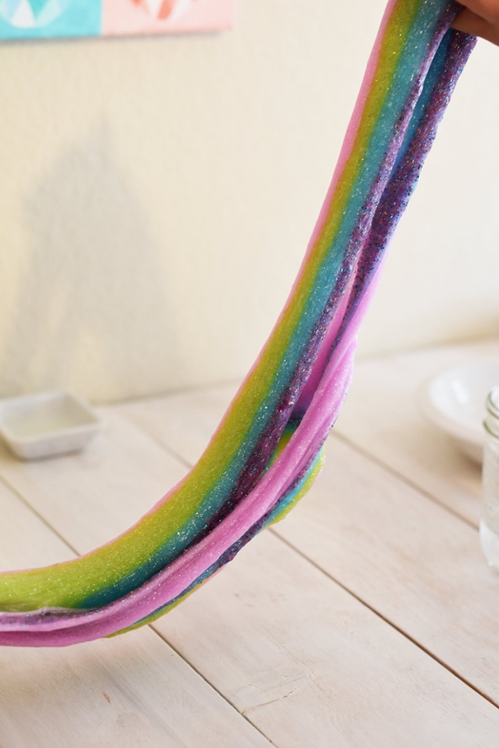 rainbow colored slime, long and decorated with glitter, being stretched over a light wooden surface, how to make slime 