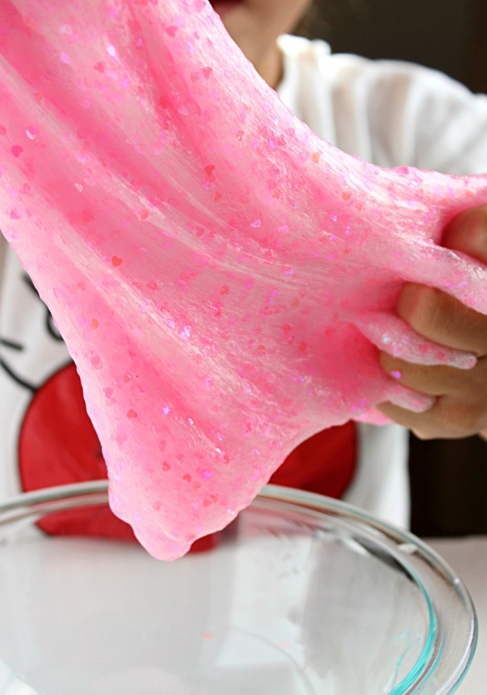 heart-shaped pink glitter flakes, inside a piece of pastel pink slime, stretched over a clear glass bowl, by a small child 