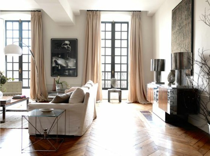 greyscale portrait framed between two tall windows, with pale beige curtains, inside a room with laminate floor, off-white sofa and other furniture
