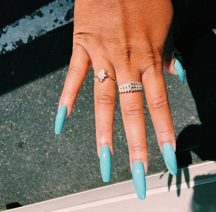 encrusted golden rings, worn on the ring and middle fingers of a tan hand, with long coffin nails, in turquoise blue