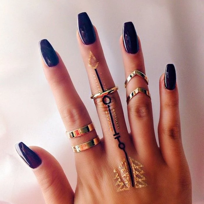 temporary hand tattoos, in black and gold, on a hand with acrylic nail shapes, painted in a smooth and glossy, dark violet nail polish