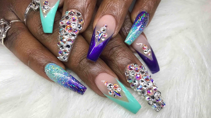 long coffin nails, painted in turquoise, purple and pale pink, and decorated with lots of rhinestones, on a pair of dark brown hands