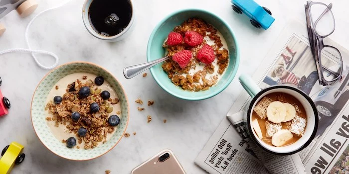 muesli and cereals, with milk and different fruit toppings, blueberries raspberries and bananas, in two bowls and a mug, on a table with lots of objects, healthy breakfast ideas