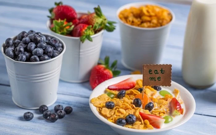 cereal with milk, covered with fruit, blueberries and grapes, sliced strawberries and clementines, healthy low calorie breakfast, berries and cornflakes in small pots nearby