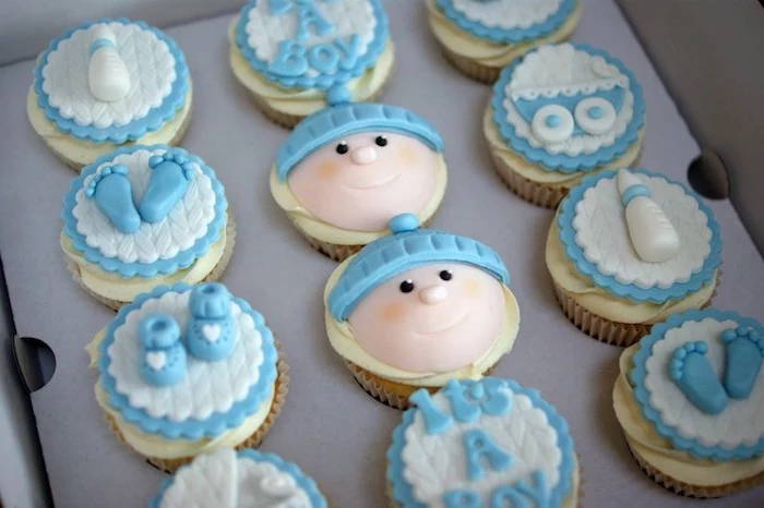 dozen of cupcakes in a cardboard box, pale yellow frosting, and blue and white fondant decorations, little feet and shoes, baby head and bottle, baby shower cakes for boys, pram and the words it's a boy