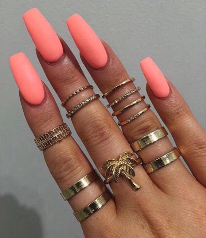 neon peach pink nail polish, on long coffin nails, attached to a tan hand, with twelve different golden rings, on a grey background