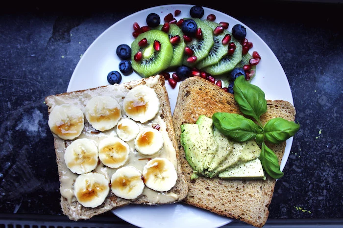 slices of bread, one with peanut butter, banana slices and honey, and one with avocado and basil, breakfast menu ideas, on a plate with kiwi slices, pomegranate seeds and blueberries