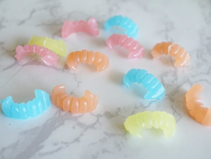 several tiny plastic vampire fangs, in different light pastel colors, yellow and orange, pink and blue, placed on a light smooth surface, with marble pattern