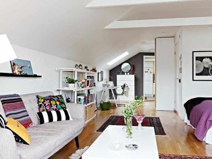 slopped ceiling in a room with white walls, containing a pale grey sofa, with multicolored cushions, white coffee table, bed and bookshelves