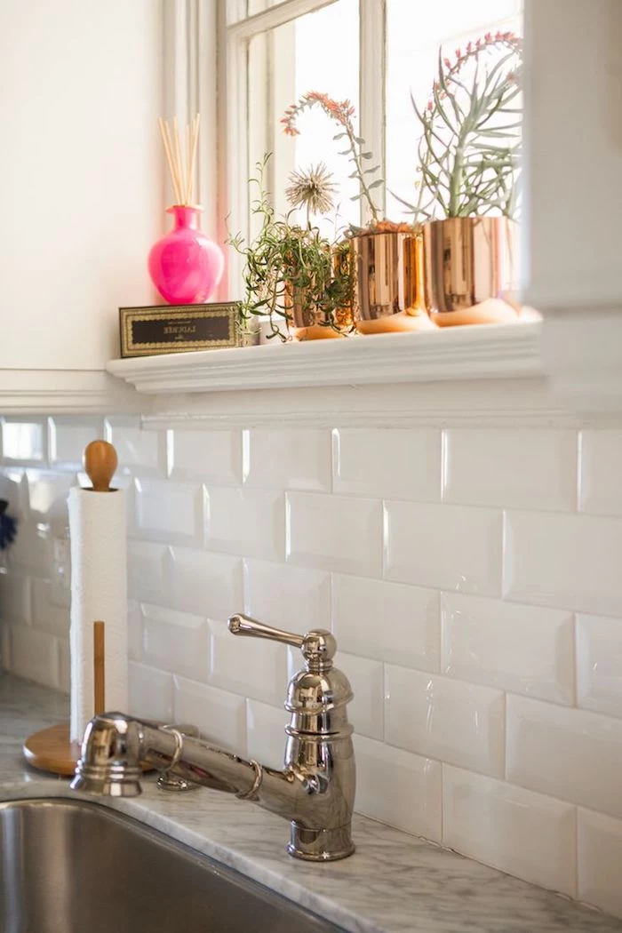 faucet in a vintage style, next to a sink and a marble counter top, white subway tile back splash, three brass planters with flowers
