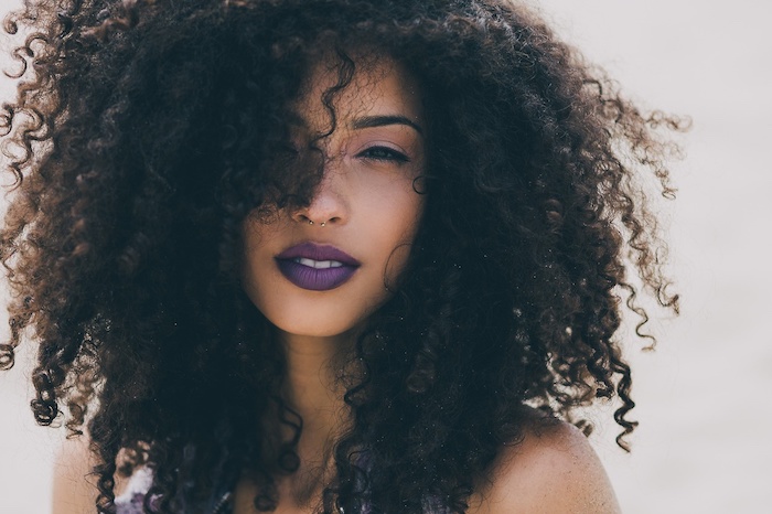 close up of a young woman, with black eyes, and dark violet lipstick, voluminous dark brunette hair, in small tight curls, hiding half of her face