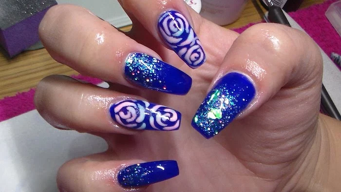 violet nail polish, decorated with pale pink and blue rose drawings, and blue iridescent glitter, on the long coffin acrylic nails, of a hand with folded fingers