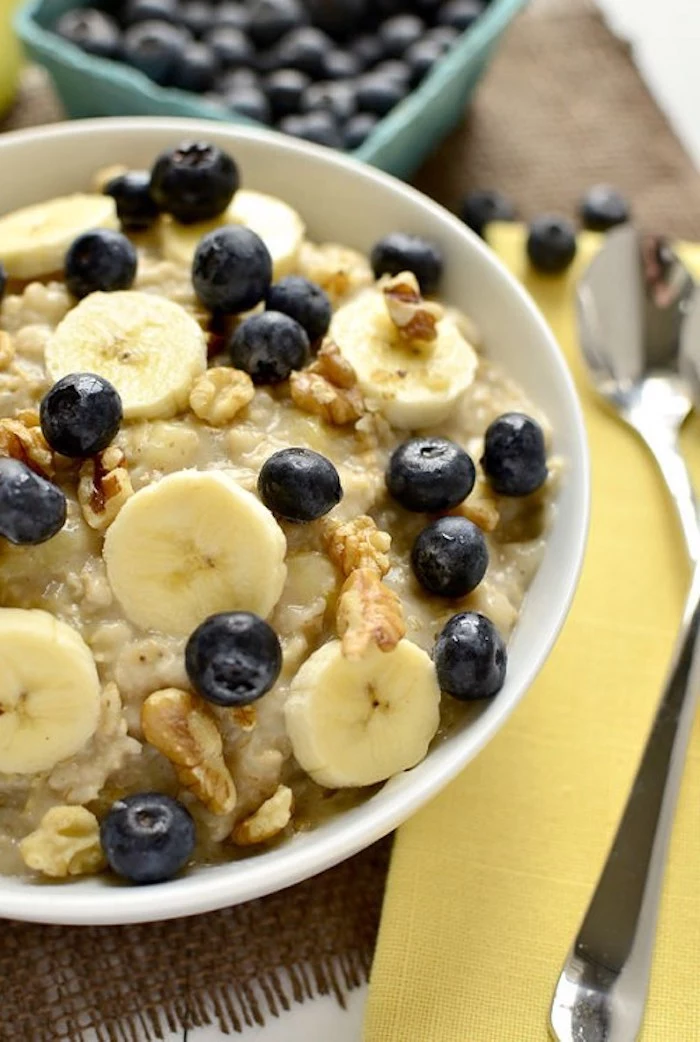 porridge topped with banana slices, blueberries and walnuts, inside a white bowl, next to a yellow paper napkin, with metal cutlery