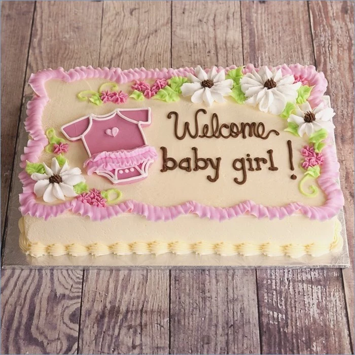 welcome baby girl, written with brown frosting, on a rectangular cake, baby shower cakes for girls, decorated with a little pink onesie, and white and pink flowers