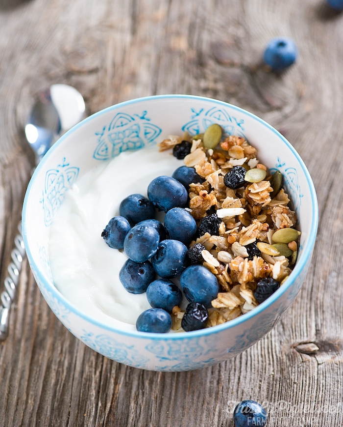 pumpkin seeds and raisins, granola and blueberries, on the side of a bowl, containing creamy yoghurt, best breakfast for weight loss, on a wooden surface 