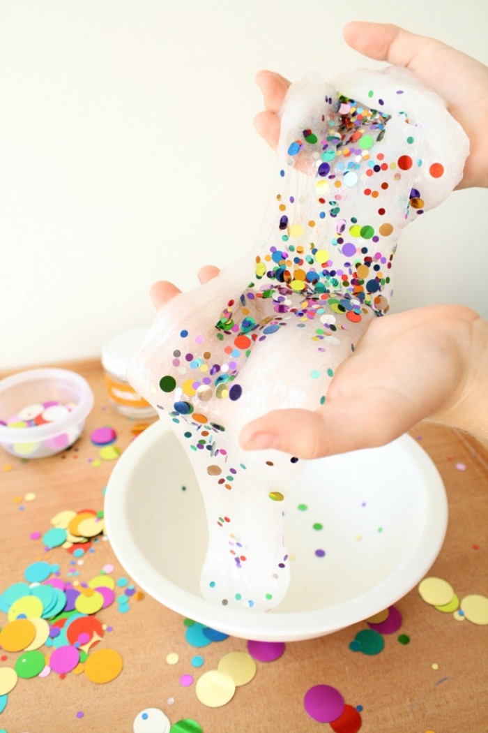 round confetti in different colors and sizes, decorating a piece of white slime, held in two pale hands, how to make slime with shaving cream, more confetti strewn about